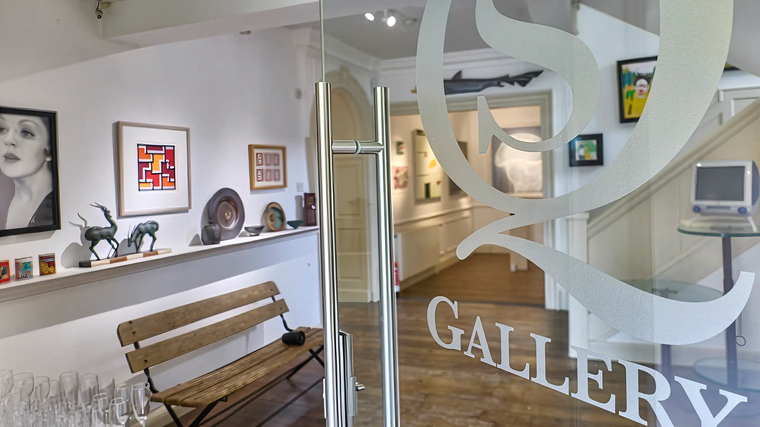 A view through the glass front doors of South Quay Gallery, showing a "GALLERY" etched logo, a wooden bench, and various artworks on white walls inside.