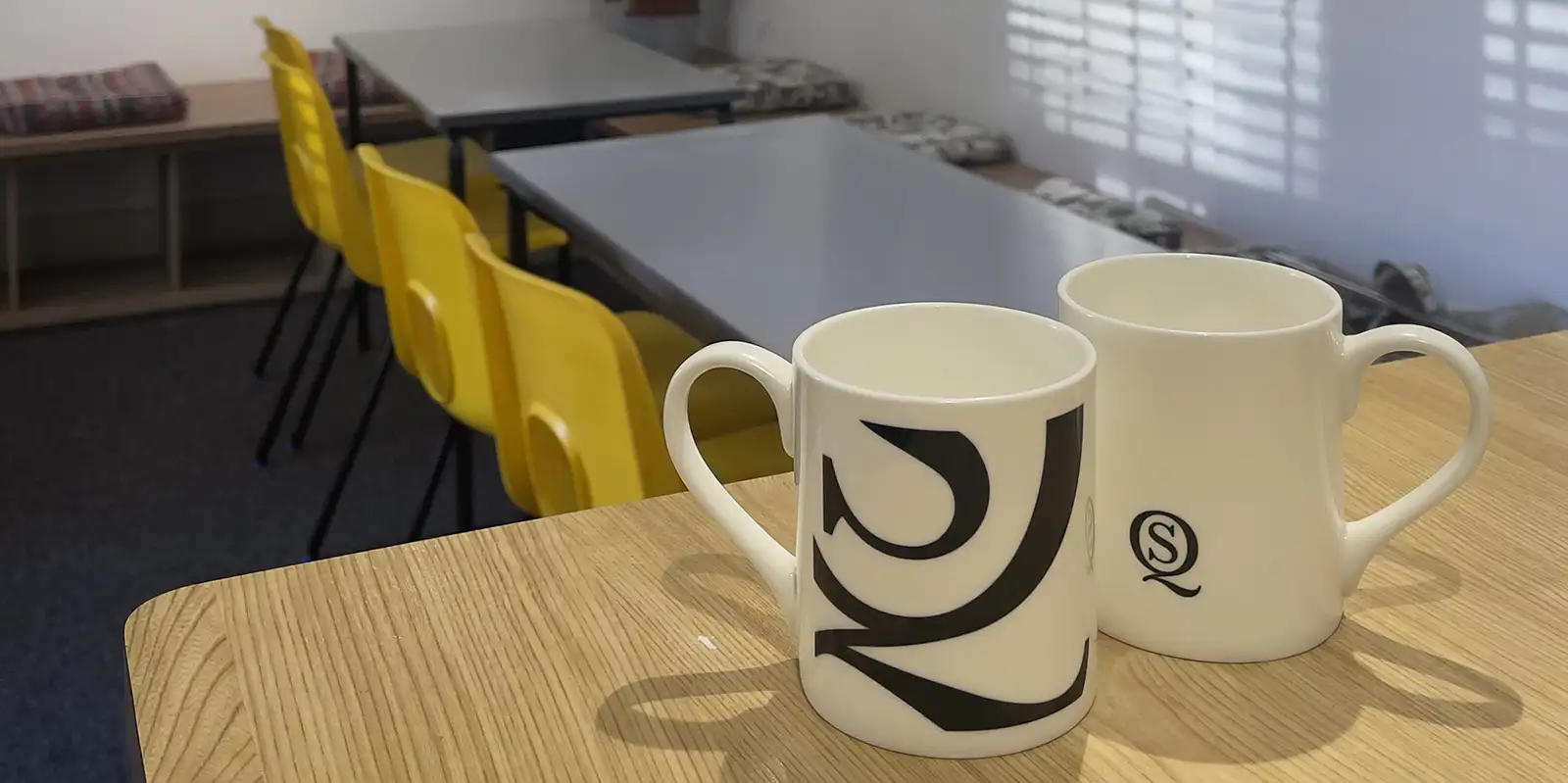 Two white mugs with the South Quay Gallery "SQ" logo on a wooden counter in the gallery cafe, Great Yarmouth.