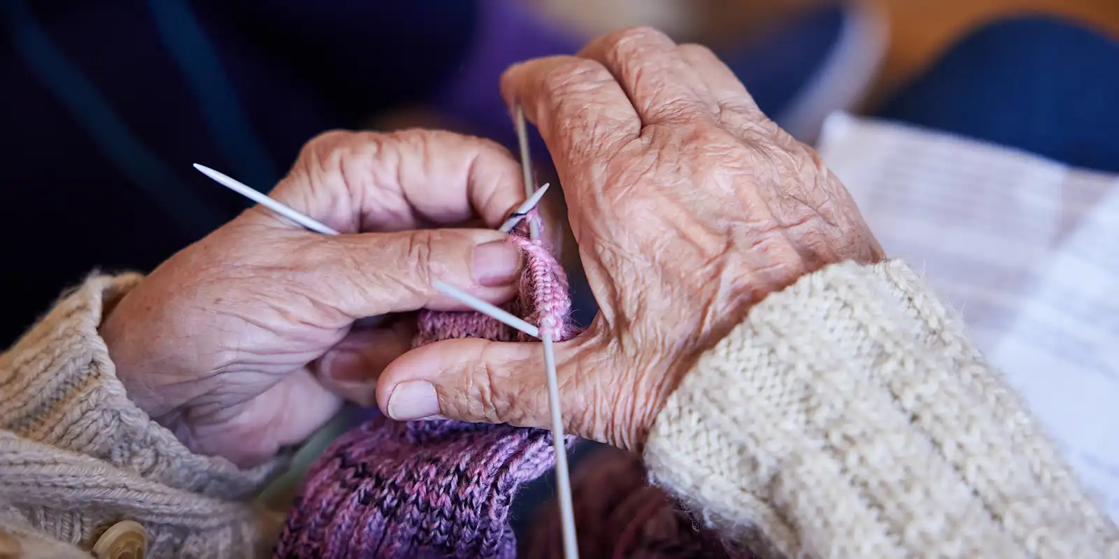 Close-up of elderly hands using silver knitting needles to knit a pink and purple patterned wool garment.