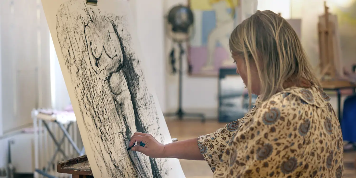 An artist in a patterned shirt works on a large-scale charcoal figure drawing during a life drawing class at South Quay Gallery.