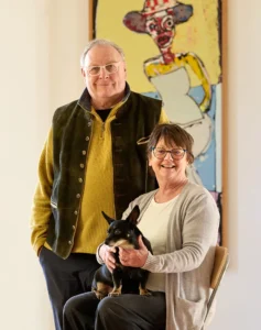 Pete and Moira Huggins standing together with their small black dog, Archie, in front of a colourful expressionist painting by John Kiki at South Quay Gallery.