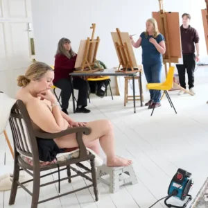 A seated life model posing in a chair with students at easels in the background in a light and airy studio
